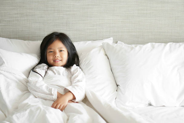 Asian child cute relax or kid girl sleep and smiling happy wake up with bright and fresh in morning on clean white bed and pillow with blanket in bedroom at home or hotel and resort on holiday - Foto, Imagem