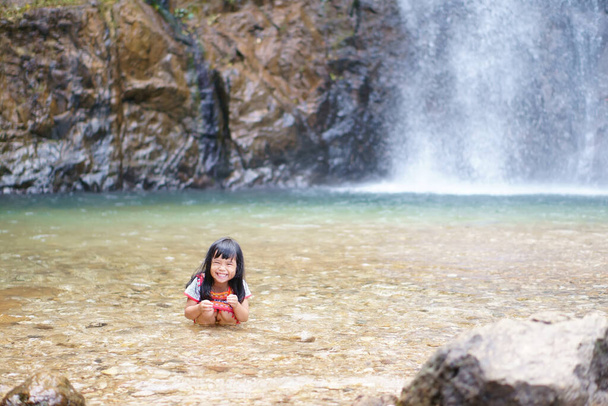 Niño asiático lindo o niña y la tribu de la colina feliz sonriendo con diversión y disfrutar jugando salpicadura de agua en la cascada o el río y vacaciones relajar viaje para viajes de verano en la cascada de Jokkradin con espacio - Foto, Imagen