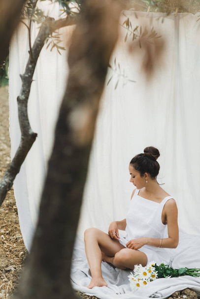 Beyaz keten elbiseli esmer kadının portresi. Arka planda asılı kumaş kumaş. Rahatlama ve rahatlama, zeytin bahçesinde piknik, bir buket papatya çiçeği.. - Fotoğraf, Görsel