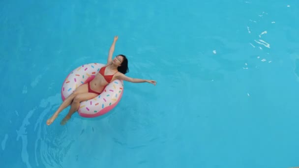 Draufsicht des schlanken jungen asiatischen Mädchens im roten Bikini-Badeanzug, das in einem aufblasbaren Rettungsring in rosa Donut liegt, die Hände hebt und im blauen Wasser schwimmt, sonniger Tag - Filmmaterial, Video