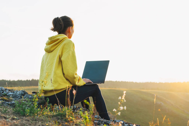 Young woman girl traveler freelancer in yellow hoodie with opened laptop looking at sunrise on beautiful view background - Фото, зображення