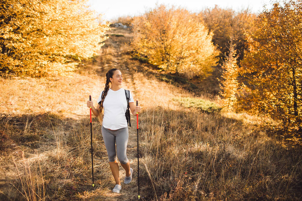 Pregnant woman nordic walking in autumn forest with backpack and trekking sticks. Healthy and active lifestyle in maternity time. Pregnancy exercises for wellness. - Photo, image