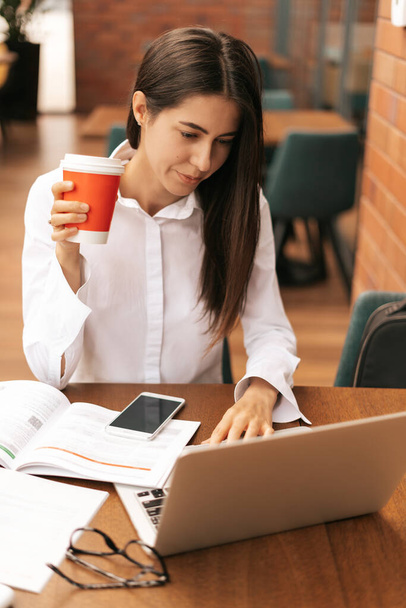 Brunette woman drinking coffee to go while working on laptop - Valokuva, kuva