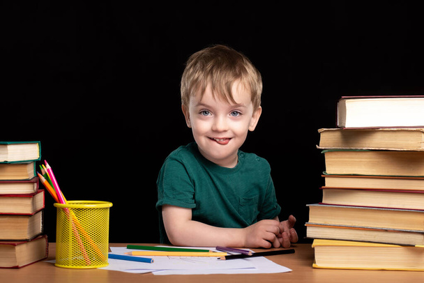 un piccolo bambino di tre anni siede a una scrivania con libri e matite contro un muro nero. apprendimento a distanza a casa. - Foto, immagini