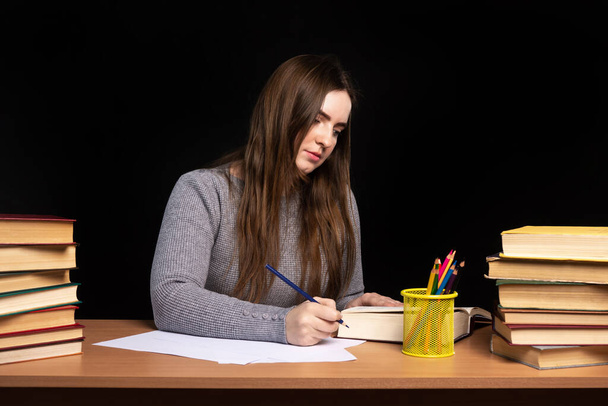 Молодая женщина студентка сидит за столом с книгами на фоне черной стены. деловая леди подписывает документы. Трудности в телекоммуникациях и обучении в карантине. - Фото, изображение