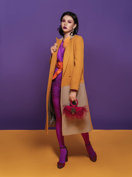 belle femme brune avec un maquillage de couleur vive dans une robe violette et une ceinture orange, avec un sac rouge et une boucle d'oreille en plume jaune est assis sur une chaise sur un fond studio violet et jaune - Photo, image