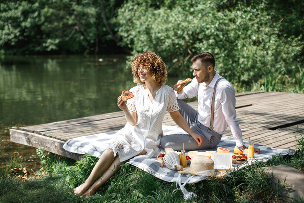 Happy couple, husband and wife are sitting in the park on a picnic and eating delicious pizza, laughing and talking. Rest in the park. Happy family concept - Foto, afbeelding