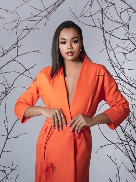 Beauty photo of young elegant african american woman.Looking at camera. Glamour makeup. Studio shot.Skin care and beauty concept.Studio shot.Happy beautiful african american woman posing in fashionable orange coat, standing on gray background - 写真・画像