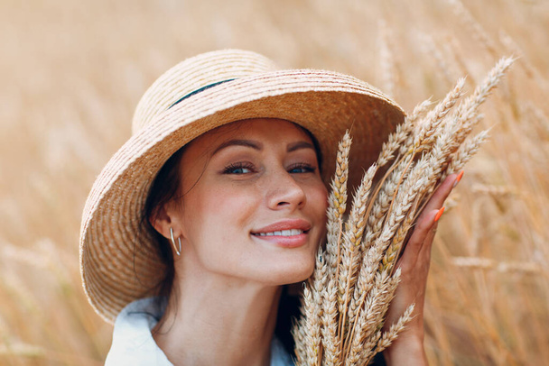 Young Woman in straw hat holding sheaf of wheat ears at agricultural field - Foto, imagen