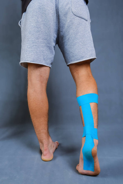 Detailní kineziologická páska nanesená na zraněné lýtko a patu bílého muže stojícího zády na modrém pozadí. Studio záběr, vertikální orientace - Fotografie, Obrázek