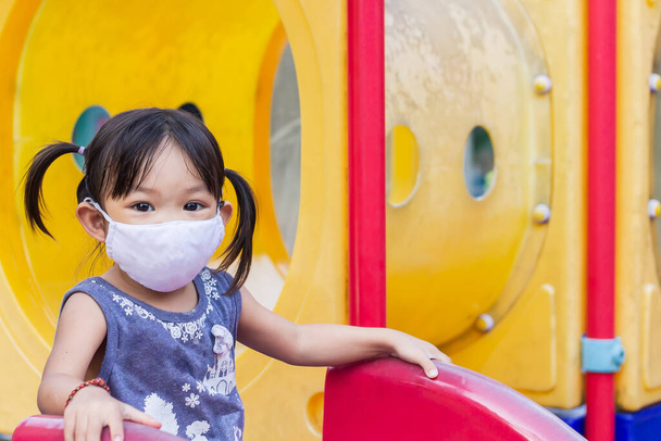 2-3 yaşlarında bir bebeğin portresi. Mutlu Asyalı kız çocuğu gülümsüyor ve kumaş maske takıyor. Çocuk parkında sürgülü bar oyuncağıyla oynuyor. Sosyal uzaklık. Çocuk konseptini öğrenmek ve aktif olmak. - Fotoğraf, Görsel