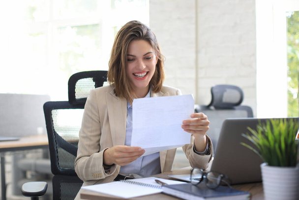 Attractive cheerful business woman checking paper documents in office, working on laptop - Фото, зображення