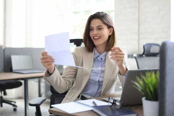 Attractive cheerful business woman checking paper documents in office, working on laptop - Foto, imagen