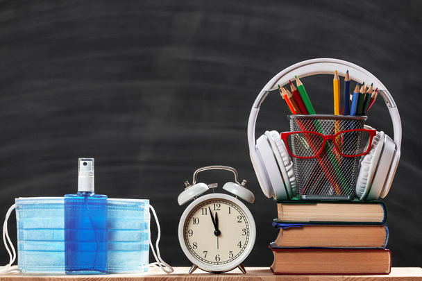 Education concept. Back to school. Schoolboy set. Stack of books pencils glasses alarm clock headphones protective medical masks sanitizer on a black board background. - Photo, Image