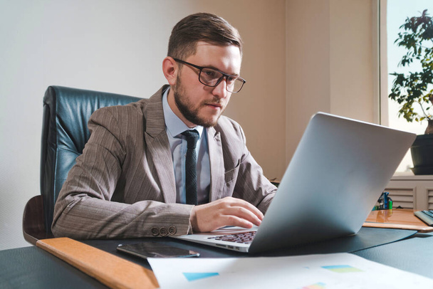Jonge zakenman in bril werkt op een laptop in zijn persoonlijke kantoor. - Foto, afbeelding
