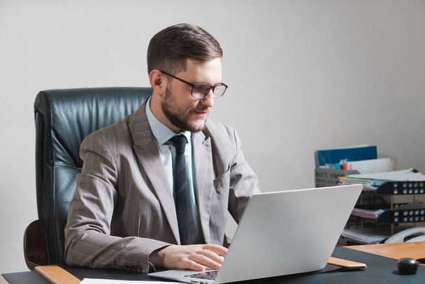 Молодой бизнесмен в очках, работающий над ноутбуком в своем личном кабинете. - Фото, изображение
