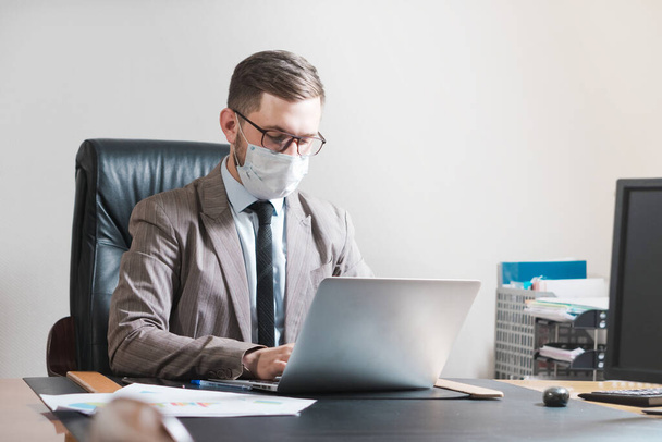 Jovem empresário de óculos e máscara facial trabalhando em um laptop em seu escritório pessoal. Negócios durante a quarentena
. - Foto, Imagem