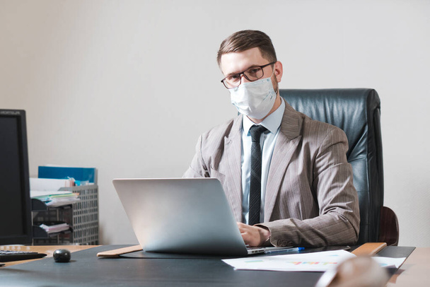 Young businessman in glasses and face mask working on a laptop in his personal office. Business during quarantine. - Foto, immagini