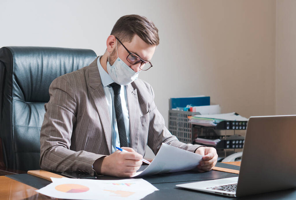 Jeune homme d'affaires en lunettes et masque facial travaillant dans son bureau personnel. Affaires en quarantaine. - Photo, image