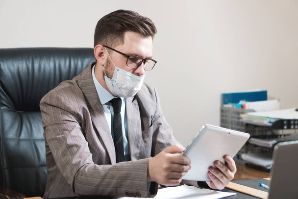 Mladý obchodník s brýlemi a obličejovou maskou pracuje na tabletu ve své kanceláři. Obchod během karantény. - Fotografie, Obrázek