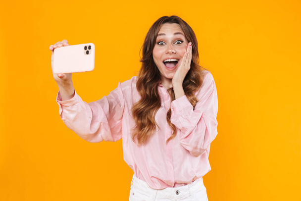 Image of excited woman taking selfie on cellphone and expressing surprise isolated over yellow background - Foto, immagini