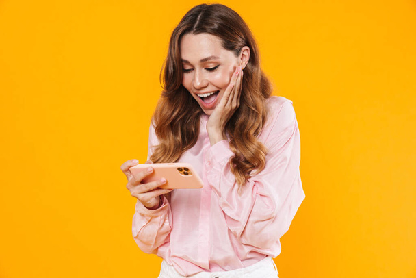 Image of cute delighted woman smiling and playing video game on cellphone isolated over yellow background - Fotó, kép