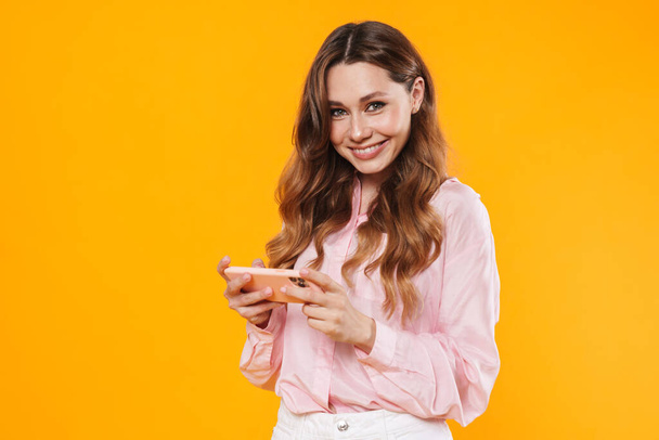 Image of beautiful pleased woman smiling and using mobile phone isolated over yellow background - Fotó, kép