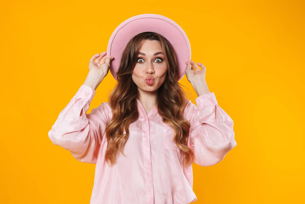 Image of attractive surprised woman in hat making kiss lips on camera isolated over yellow background - Zdjęcie, obraz