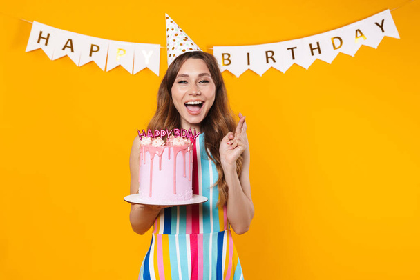 Image of happy woman in party cone holding torte and fingers crossed for good luck isolated over yellow background - Foto, Bild