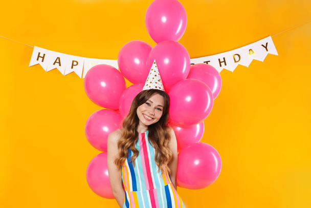Image of cheerful young woman in party cone holding pink balloons and smiling isolated over yellow background - Zdjęcie, obraz