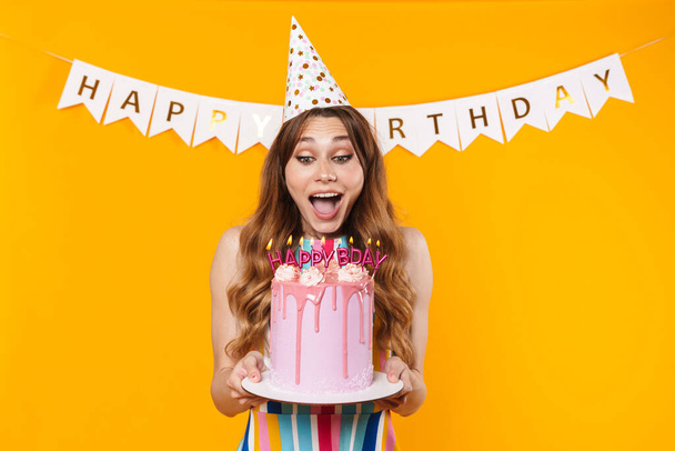 Image of delighted young woman in party cone showing birthday torte with candles isolated over yellow background - Фото, зображення