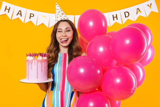 Image of excited cute woman in party cone posing with pink balloons and birthday torte isolated over yellow background - Photo, Image
