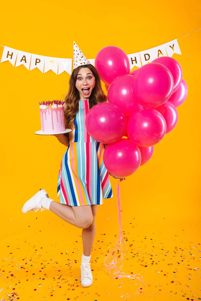 Image of excited cute woman in party cone posing with pink balloons and birthday torte isolated over yellow background - Photo, image