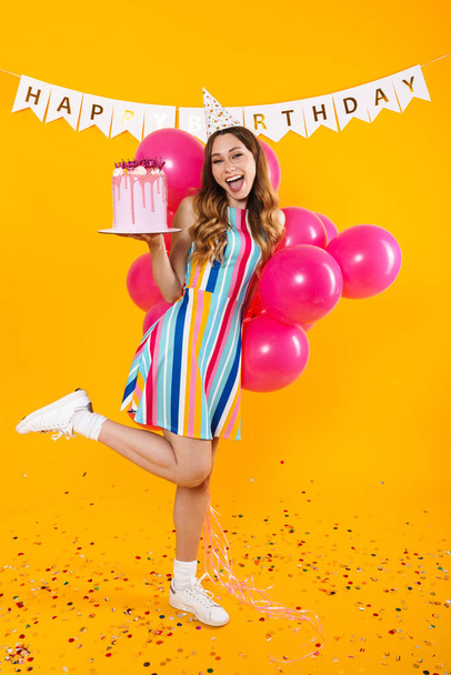 Image of excited cute woman in party cone posing with pink balloons and birthday torte isolated over yellow background - Fotó, kép