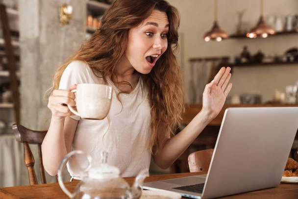 Beautiful young smiling girl sitting at the kitchen table, using laptop computer, video chatting, drinking coffee - 写真・画像