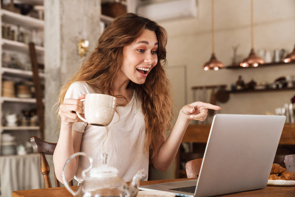 Mooi jong lachend meisje zitten aan de keukentafel, met behulp van laptop computer, video chatten, het drinken van koffie - Foto, afbeelding