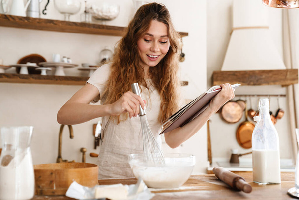 Attractive young cheerful girl baking at the kitchen, making dough, holding recipe book, having ideas - Φωτογραφία, εικόνα