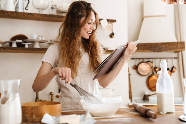 Attractive young cheerful girl baking at the kitchen, making dough, holding recipe book, having ideas - Valokuva, kuva