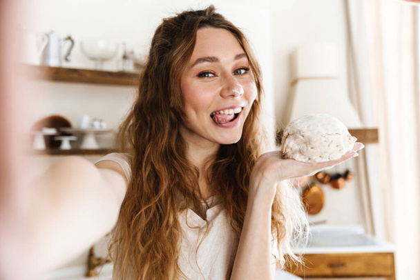Emocionado joven encantadora chica tomando una selfie mientras hornear en la cocina, mostrando la masa - Foto, Imagen