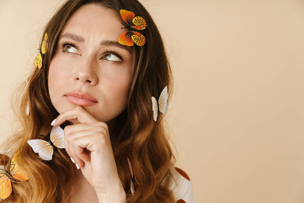 Photo of beautiful perplexed woman with fake butterflies posing and looking aside isolated over beige wall - Photo, Image