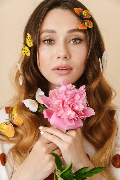 Photo of beautiful young woman with fake butterflies and peony looking at camera isolated over beige wall - Photo, Image
