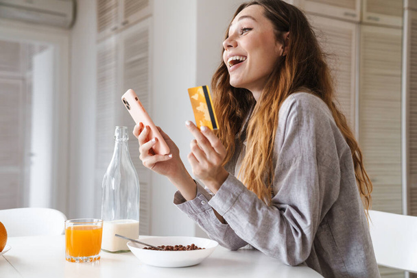 Pretty cheerful young woman having breakfast while sitting at the kitchen table, using mobile phone and credit card for shopping online - Foto, Imagen