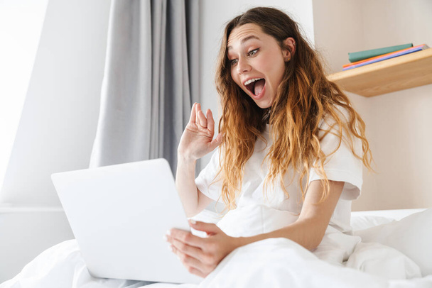 Portrait of ginger excited woman waving hand and using laptop while sitting in bed after sleep - Фото, изображение