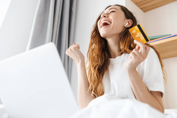 Portrét vzrušené ženy s gestem vítěze sluchátka a držení kreditní karty při použití notebooku v posteli - Fotografie, Obrázek