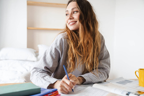 Fénykép mosolygós gyönyörű diák nő írás a füzetbe, miközben a házi feladatot a hálószobában - Fotó, kép