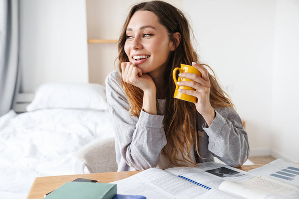 Photo of smiling beautiful student woman drinking tea while doing homework in bedroom - Foto, imagen