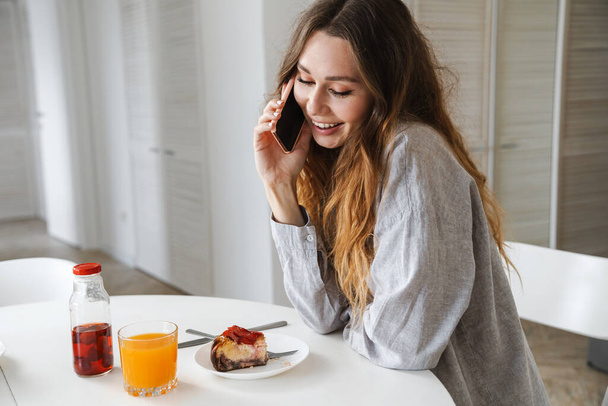 Photo of funny beautiful woman laughing and talking on cellphone while having breakfast in white kitchen - Photo, Image