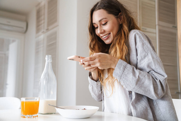 Image of cheerful beautiful woman taking photo on smartphone while having breakfast in white kitchen - Foto, afbeelding
