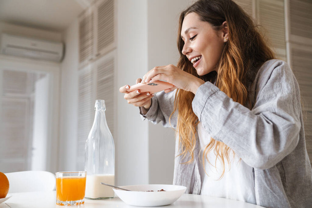Image of cheerful beautiful woman taking photo on smartphone while having breakfast in white kitchen - Фото, зображення