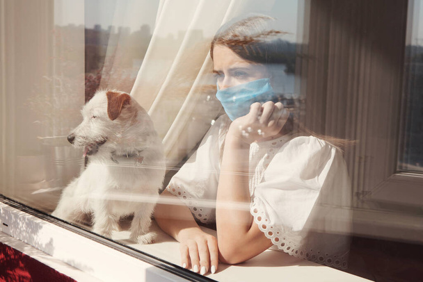 Smutná mladá žena v lékařské masce se psem Jackem Russellem Broken zůstat v izolaci doma pro vlastní karanténu. Koncepce domácí karantény, prevence COVID-19, nákaza koronavirem - Fotografie, Obrázek